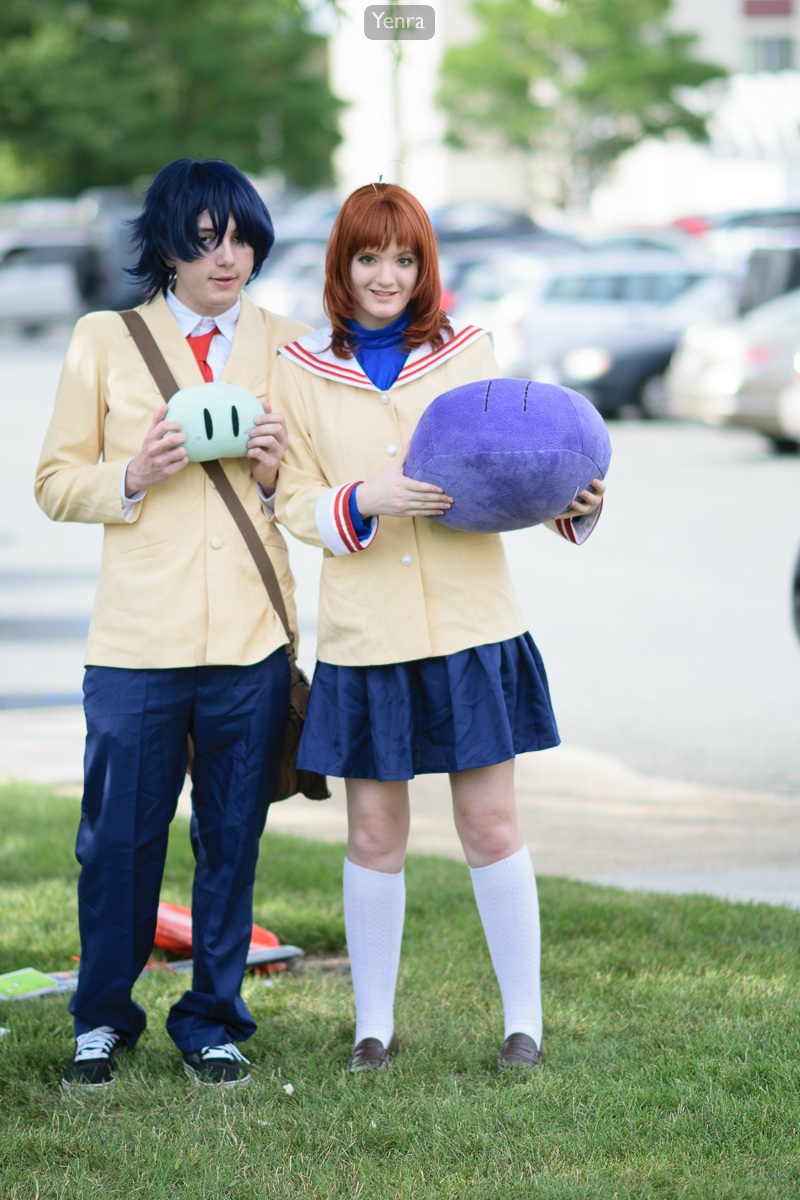 Two individuals in school uniforms pose with masks and toys on a grassy area near parked cars.