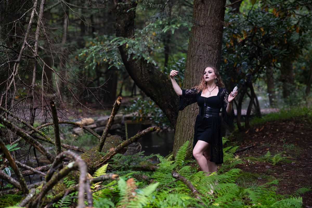 A woman stands in a forest clearing, surrounded by trees and foliage, holding a crystal or stone.