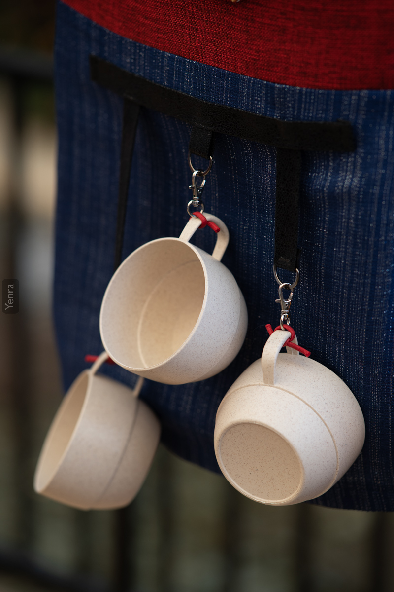 A person wearing a blue apron with red trim and four white mugs suspended from it.