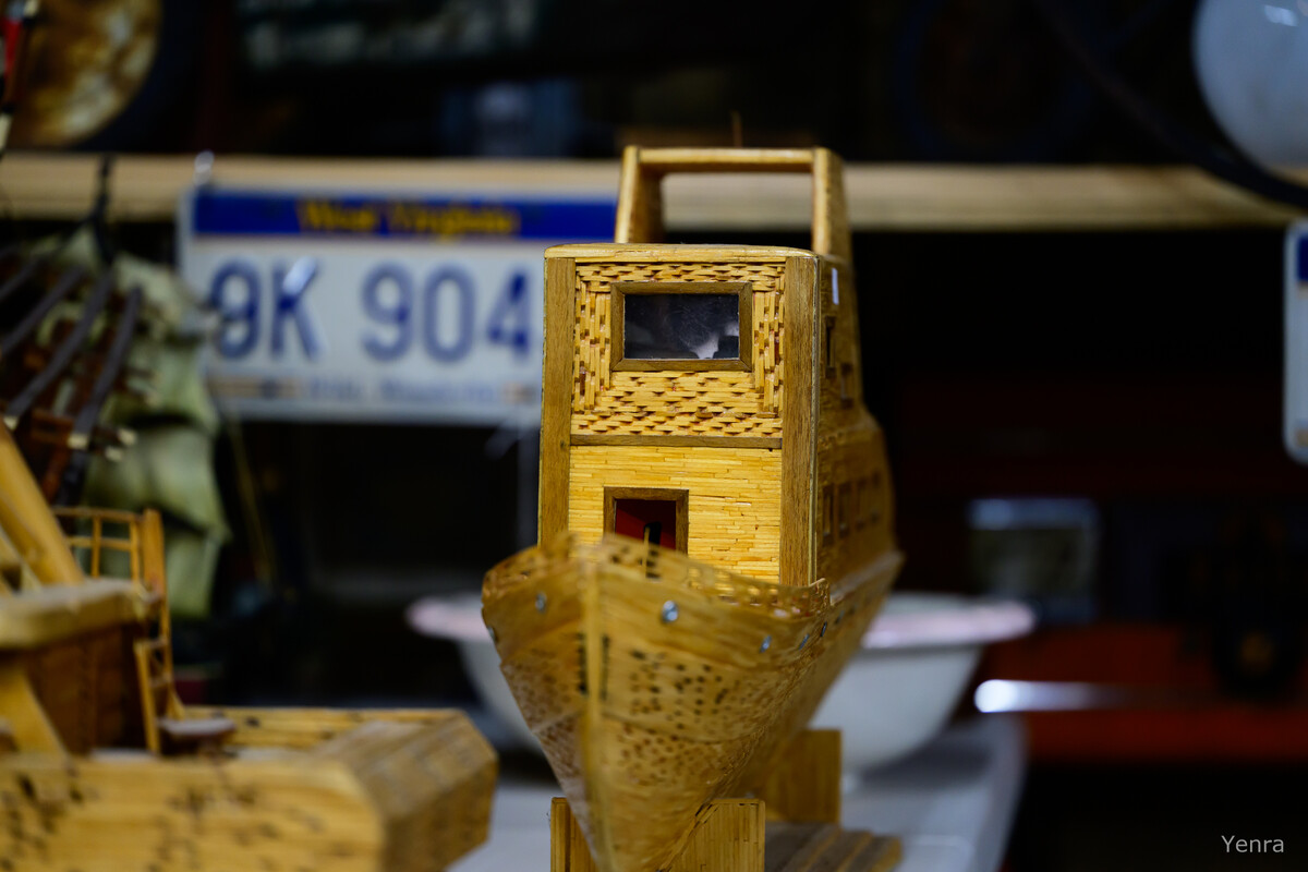 Close-up of a woven bamboo model boat with a cabin and handle, sitting in front of a blurred license plate.