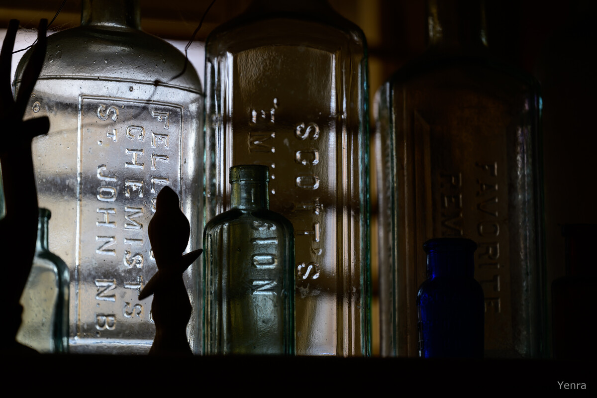 Backlit antique glass bottles with embossed text, framed by dark silhouettes of standing figures in low light.