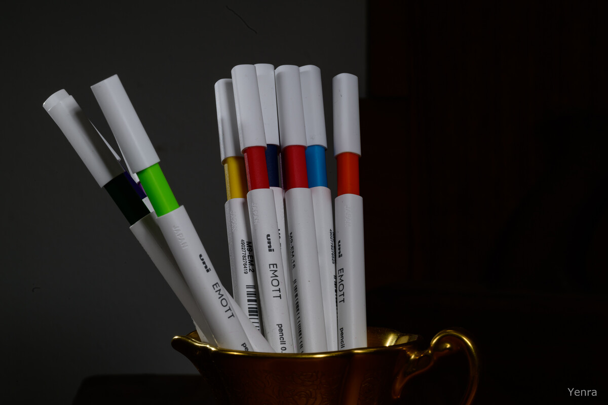 Several Uni Emott colored pencils with white barrels and colorful eraser tips (green, yellow, red, blue, orange) are arranged upright in a decorative gold cup against a dark background.