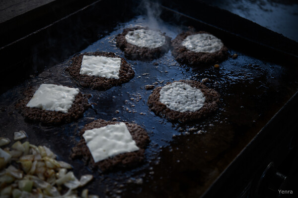 Six burger patties with melting cheese cooking on a hot griddle next to chopped onions
