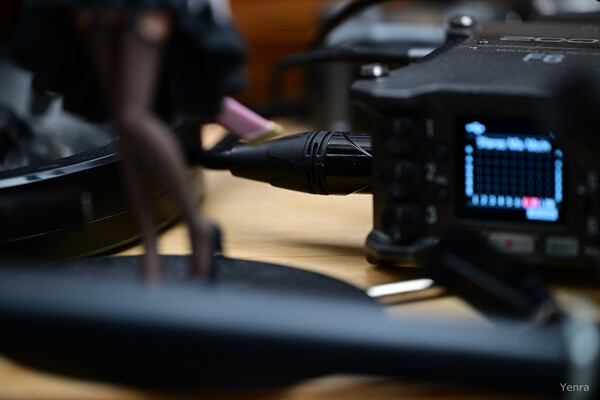 Close-up of a Zoom F6 audio recorder connected by cable, with a blurred figure in the foreground on a wooden surface.