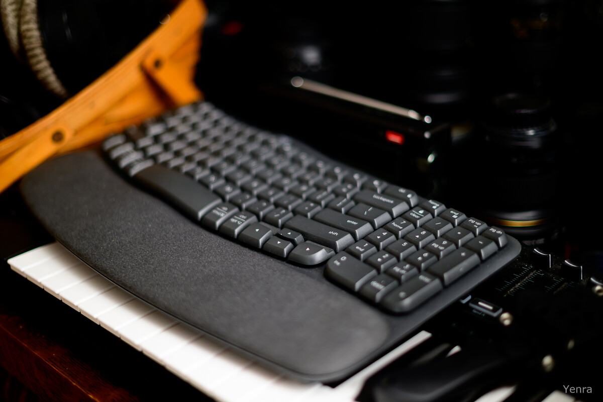 Close-up of a black Wave wireless keyboard with wrist rest, placed on white keys next to camera lenses and a wooden tool.